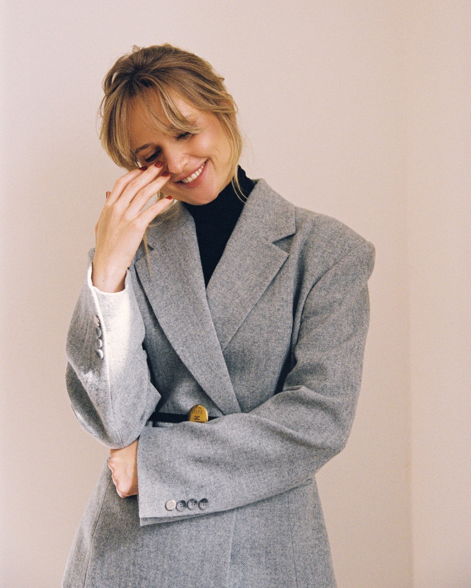 A woman wearing a wide, light gray wool-blend blazer over a black turtleneck, posing against a solid pale beige background.