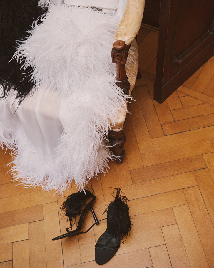 White silk chiffon bridal cape with natural ostrich feather trim draped over a vintage chair, with black feather-trimmed high-heeled sandals on the wooden parquet floor.