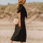 Woman in a black dress holding a round beige bag on a beach.