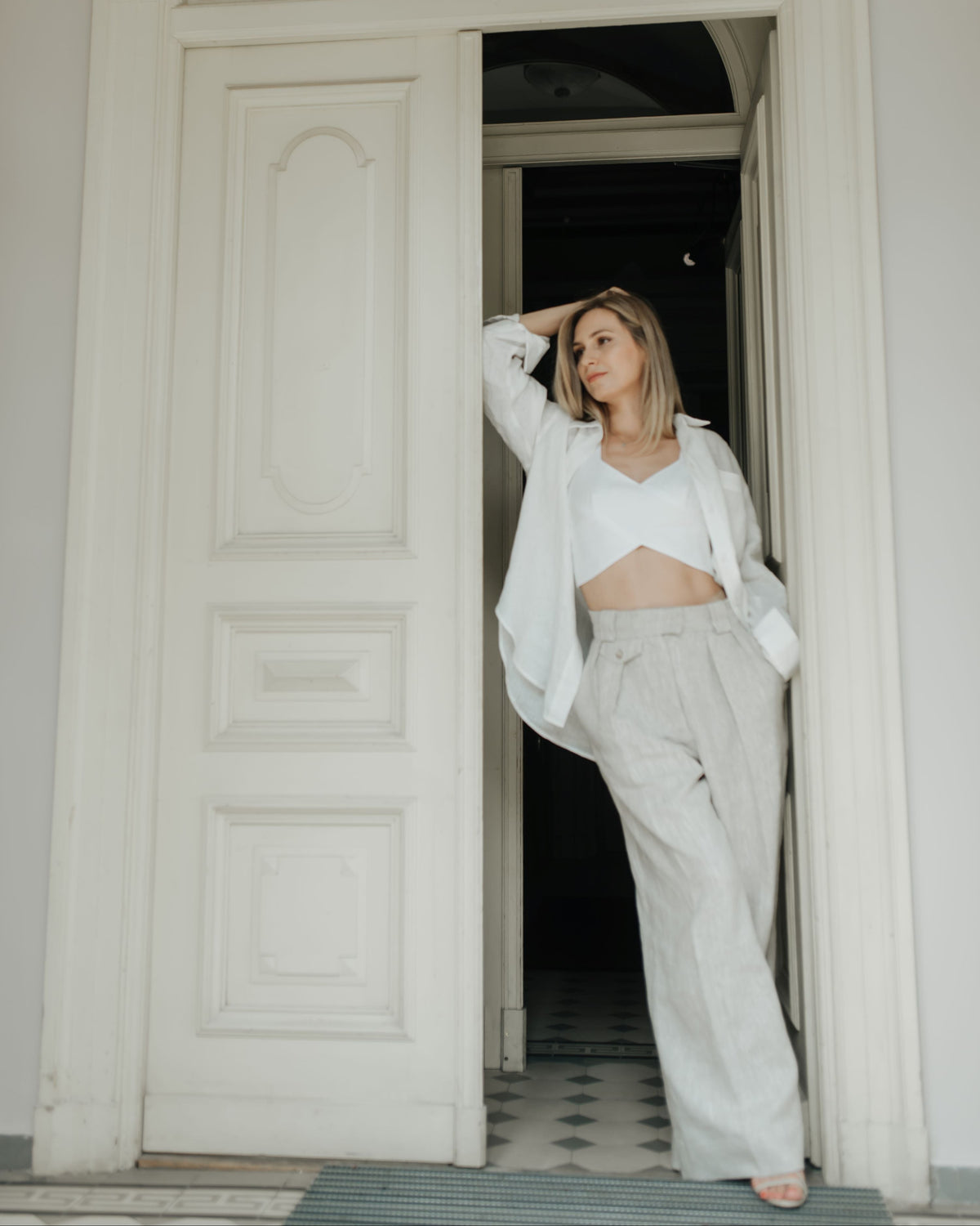A woman posing in a doorway wearing a white cropped tank top, a loose white button-up shirt, and natural linen wide-leg trousers.