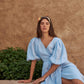 A woman wearing a sky blue puff-sleeve wrap dress sits on a stone ledge against a textured brown stucco wall.