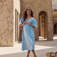 A woman wearing a light blue midi dress with puff sleeves and a V-neck stands in a desert-style courtyard with beige adobe buildings. A woven straw bag is on the ground next to her.