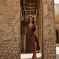 A woman wearing a brown wrap-style midi dress in a rustic stone archway doorway.