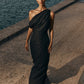 Woman in a black one-shoulder dress standing by a waterfront.