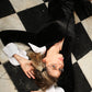 Woman in a black velvet midi dress with detachable white flared cuffs, lying on a black-and-white tiled floor.