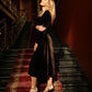Woman standing on a staircase in a brown velvet midi dress with a fitted waist and flared skirt.