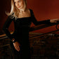 Woman wearing a brown velvet dress with long sleeves and a square neckline, shown from the front beside an ornate railing.