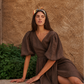 A woman wearing a brown puff-sleeve midi dress and a patterned headband poses on a stone ledge against a textured stucco wall.