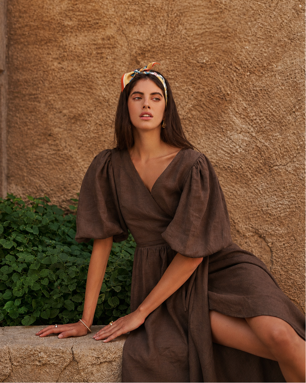 A woman wearing a brown puff-sleeve midi dress and a patterned headband poses on a stone ledge against a textured stucco wall.