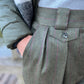 Close-up of a person wearing pleated wool trousers in dark green with thin pink pinstripes, paired with a gray knit top and an olive puffer jacket.
