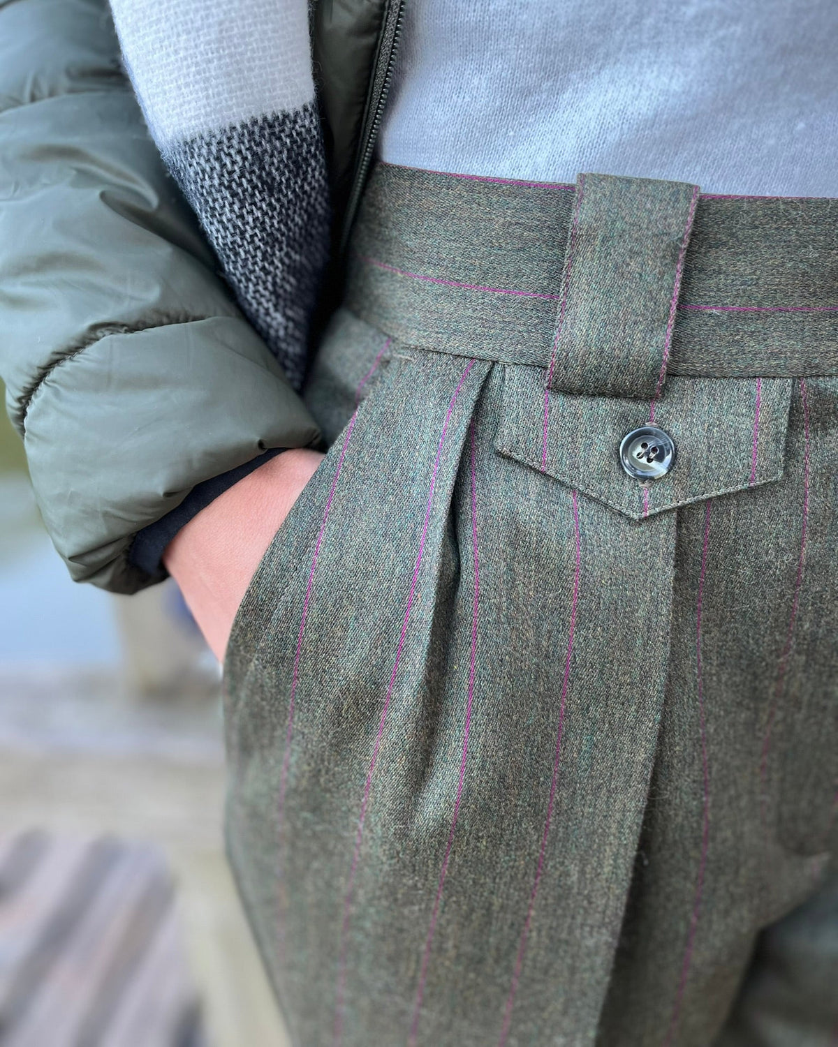 Close-up of a person wearing pleated wool trousers in dark green with thin pink pinstripes, paired with a gray knit top and an olive puffer jacket.