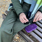A person sitting on a wooden park bench outdoors, wearing an olive green puffer jacket and matching trousers, with a multicolor scarf and black sunglasses resting on their lap. A purple velvet case is placed on the bench beside them.