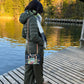A person standing on a wooden dock by a lake, wearing an olive green wide-leg pants, a light scarf, and carrying a small dark crossbody bag with colorful floral embroidery and a gold-tone logo.