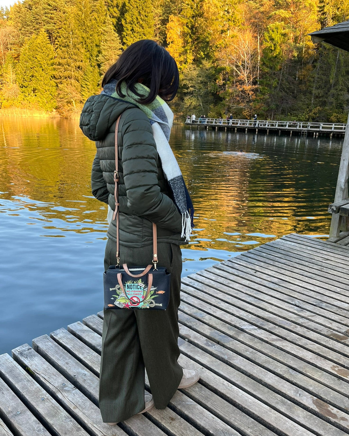 A person standing on a wooden dock by a lake, wearing an olive green wide-leg pants, a light scarf, and carrying a small dark crossbody bag with colorful floral embroidery and a gold-tone logo.