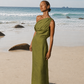 Woman wearing a green one shoulder linen dress with soft draped neckline and fitted waist, standing barefoot on a sandy beach by the ocean. The dress has an ankle-length silhouette and relaxed draping across the bust.