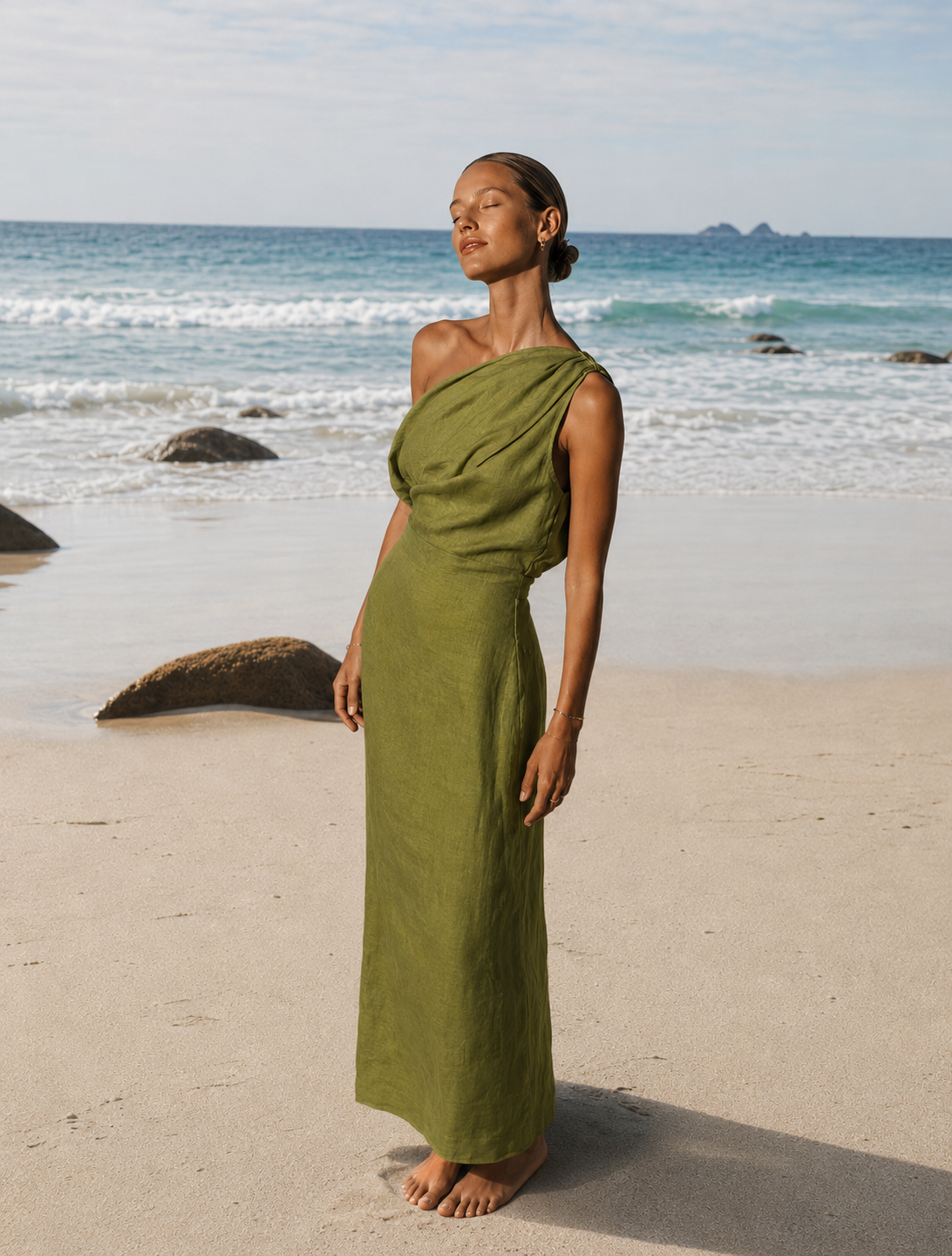 Woman wearing a green one shoulder linen dress with soft draped neckline and fitted waist, standing barefoot on a sandy beach by the ocean. The dress has an ankle-length silhouette and relaxed draping across the bust.