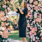 Woman in a black dress standing between two floral arches with pink and white flowers.