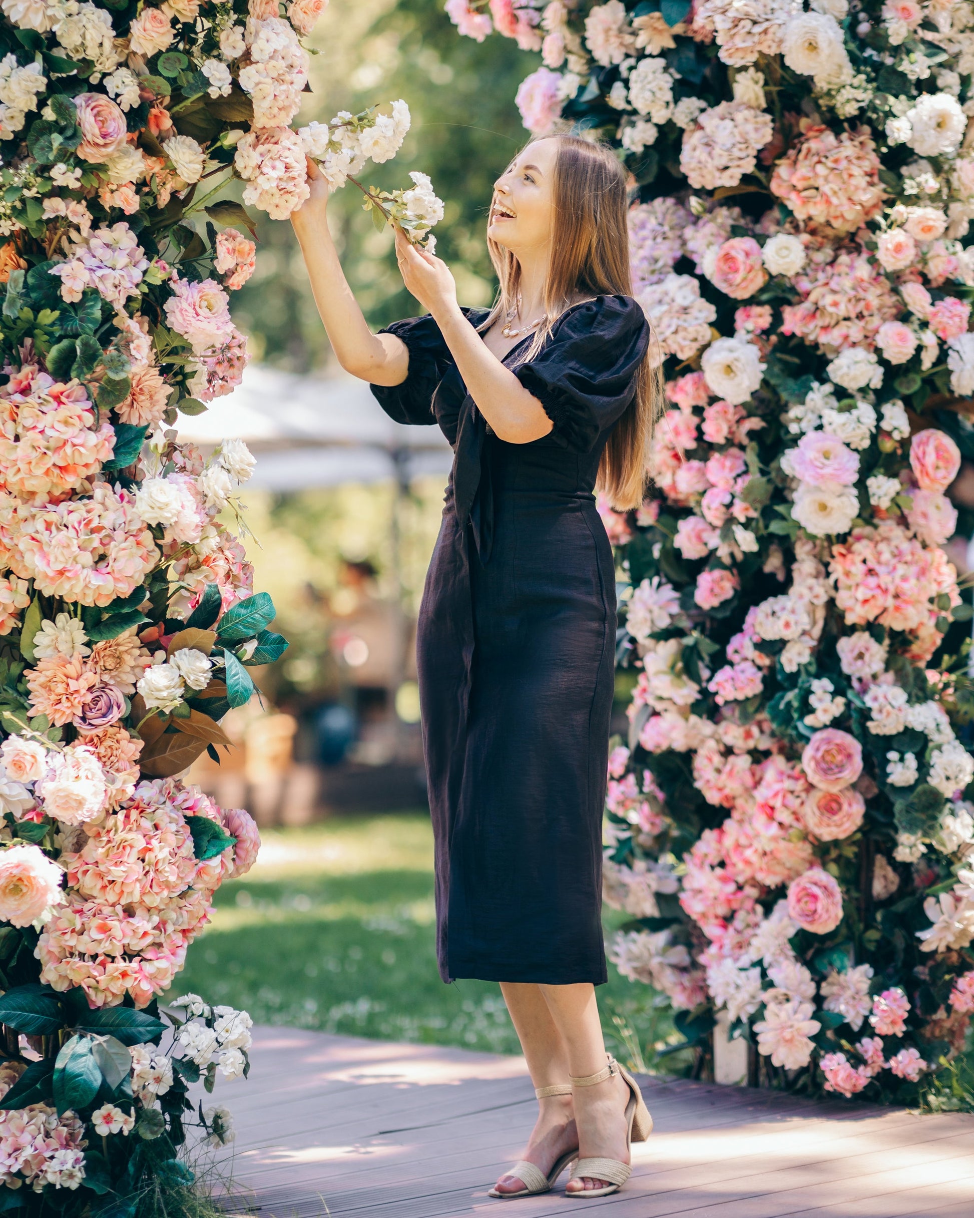 Woman in a black dress standing between two floral arches with pink and white flowers.