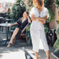 Woman in a white outfit standing outdoors with greenery and people in the background