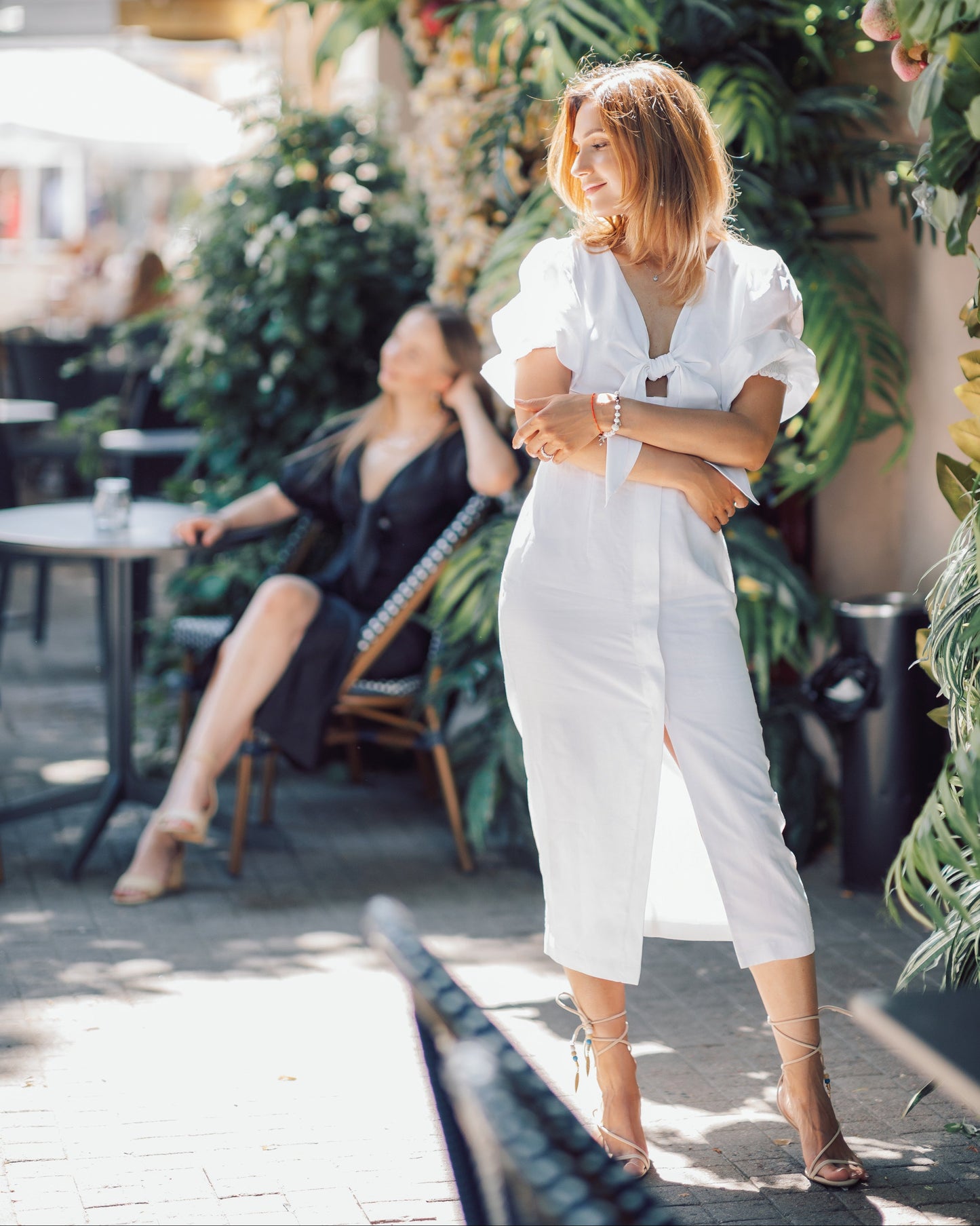 Woman in a white outfit standing outdoors with greenery and people in the background