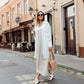 Woman in a white outfit walking on a street with buildings and outdoor seating in the background.