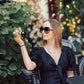 Woman in a black dress and sunglasses sitting outdoors with plants and flowers in the background