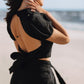 Close-up of the LUNA black linen top with a dramatic open back and bow tie, styled with black linen trousers by the sea.