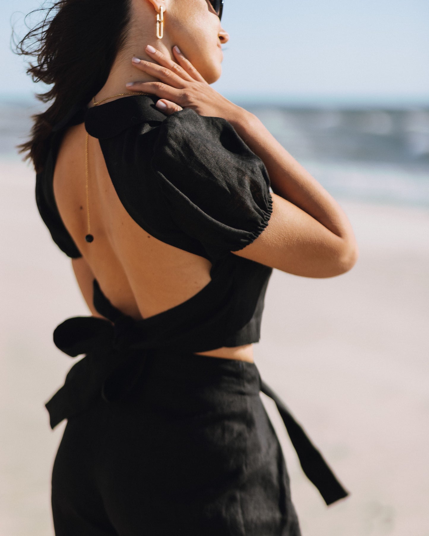 Close-up of the LUNA black linen top with a dramatic open back and bow tie, styled with black linen trousers by the sea.