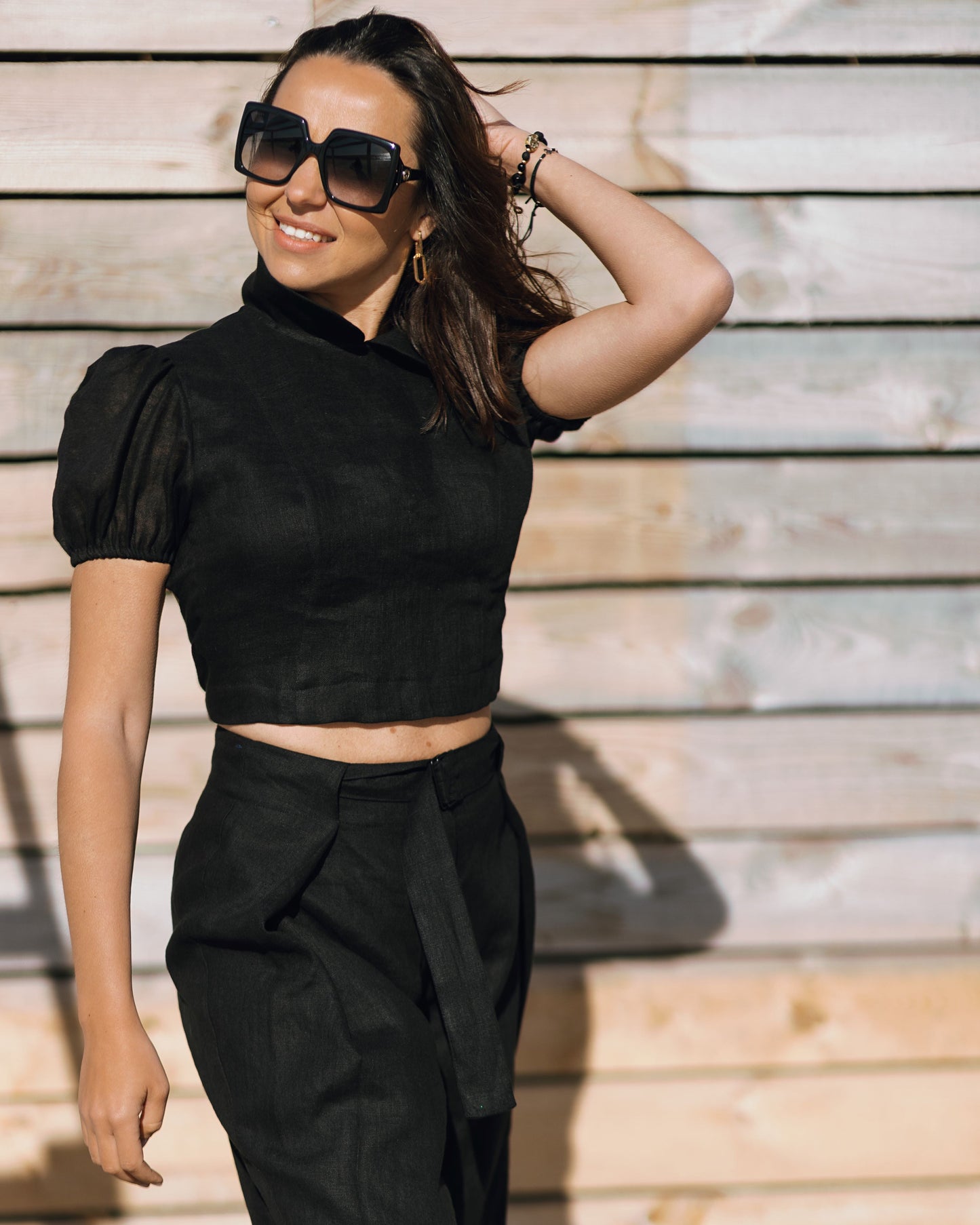 Woman in the LUNA black linen crop top with puff sleeves and a high neckline, styled with matching black trousers in warm sunlight.