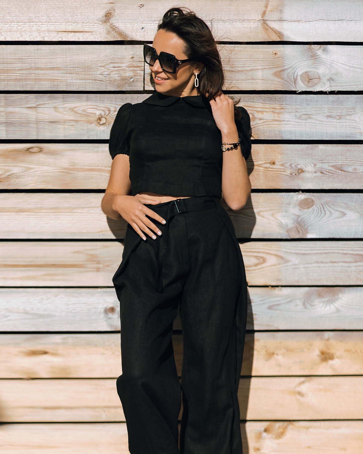Woman wearing the LUNA black linen crop top with matching wide-leg linen trousers, creating a minimal all-black summer outfit in the sunlight.