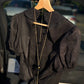 LUNA black linen top on a wooden hanger in warm sunlight, showing the structured collar, puff sleeves and natural texture of the linen fabric.