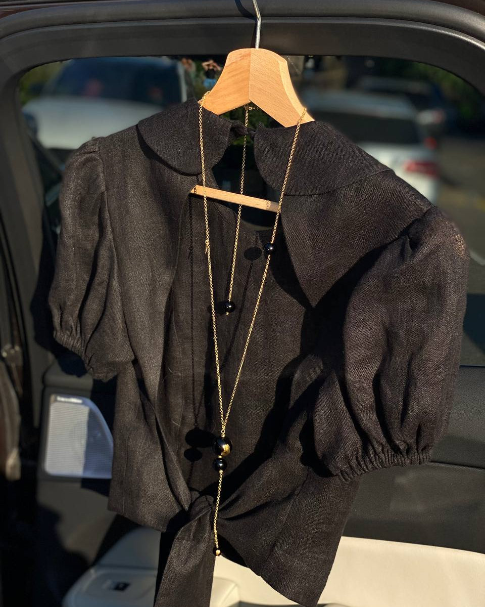 LUNA black linen top on a wooden hanger in warm sunlight, showing the structured collar, puff sleeves and natural texture of the linen fabric.