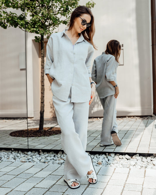 A woman wearing a light grey linen suit with a relaxed fit. The suit includes a shirt with a classic collar and 3/4 sleeves, and trousers with a high-rise waist and side pockets.