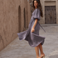 A woman wearing a flowing lavender midi dress with puff sleeves and a tie waist, posing in an outdoor alleyway with textured beige walls.
