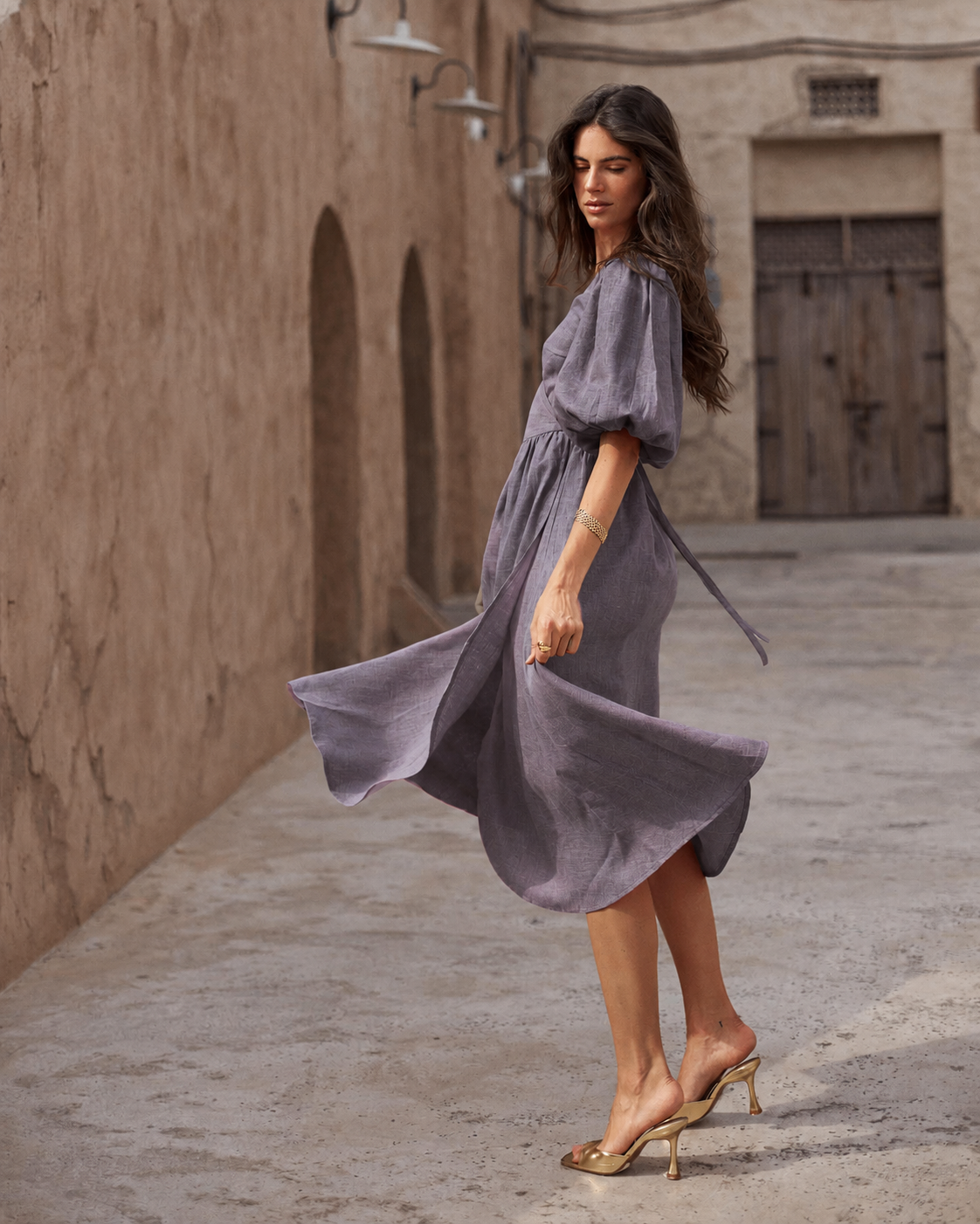 A woman wearing a flowing lavender midi dress with puff sleeves and a tie waist, posing in an outdoor alleyway with textured beige walls.