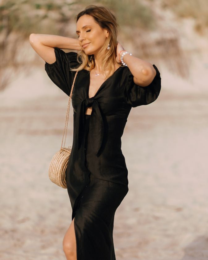 A woman standing on a beach wearing a black midi dress with puff sleeves and a knot front detail.