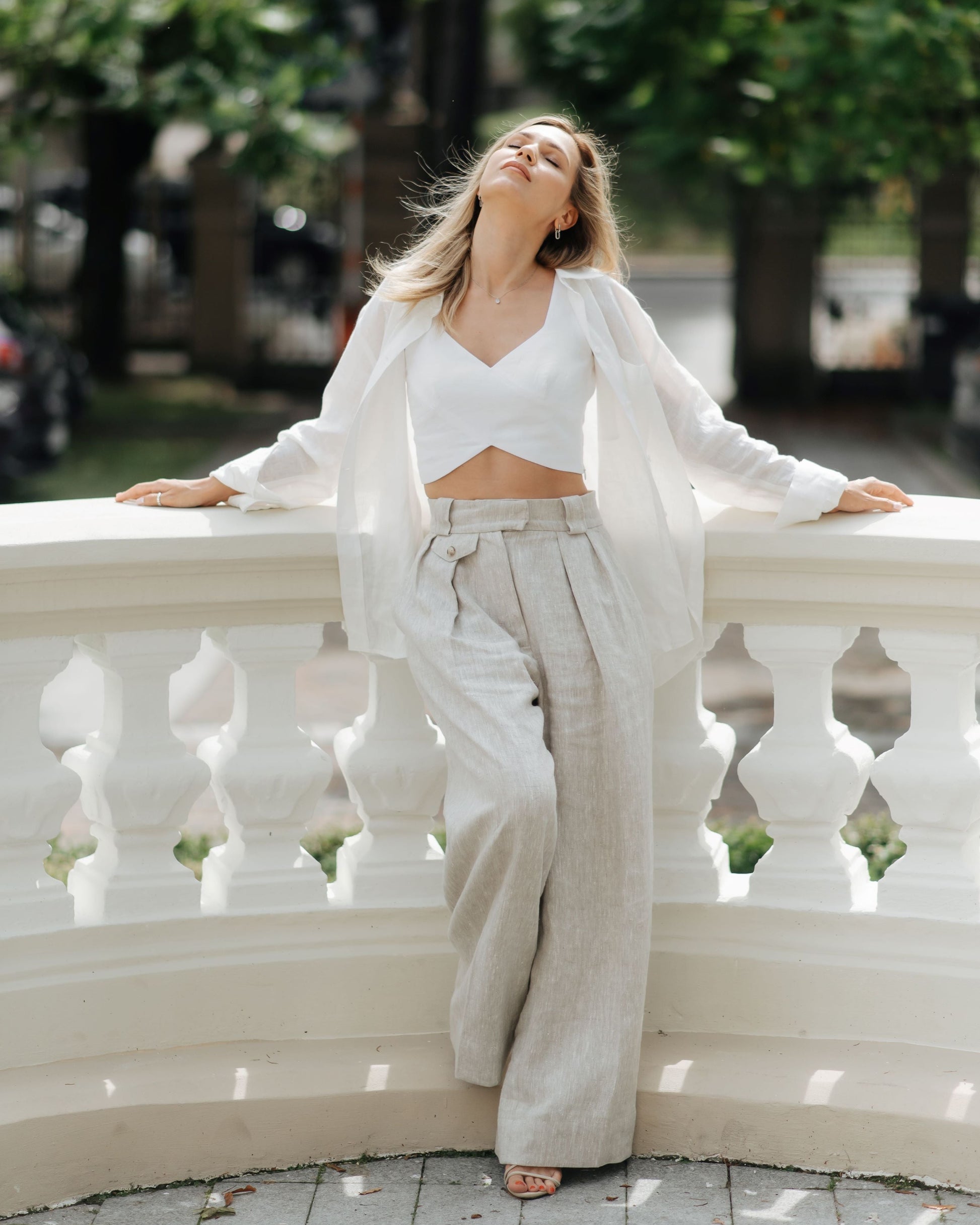 A woman seated on a balcony wearing high waisted beige linen trousers and a white top, with trees and buildings in the background.