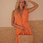A person wearing an orange sleeveless linen vest with front button closure and pockets, paired with a white top, and holding a straw bag.