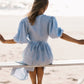 A woman seated on a bench at the beach, wearing a blue wrap dress with puff sleeves and a V-neckline, with the ocean in the background.