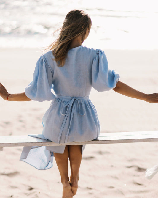 A woman seated on a bench at the beach, wearing a blue wrap dress with puff sleeves and a V-neckline, with the ocean in the background.