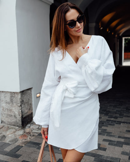 A woman wearing a white linen kimono dress with long sleeves, a V-neckline, and elastic cuffs. The dress features a kimono style front wrap and an asymmetrical bottom.