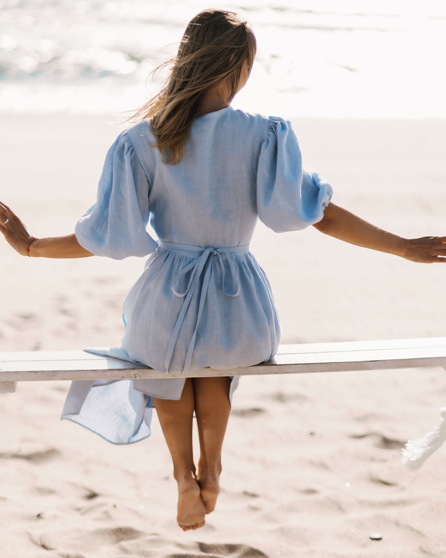 A woman sitting on a white beach chair on a sandy beach, viewed from behind. She is wearing a sky blue linen dress with puffed sleeves and a tie waist.