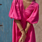 A woman wearing a fuchsia pink midi dress with voluminous puff sleeves and a wrap-style V-neckline, standing against a rustic blue wooden door and beige stucco wall.