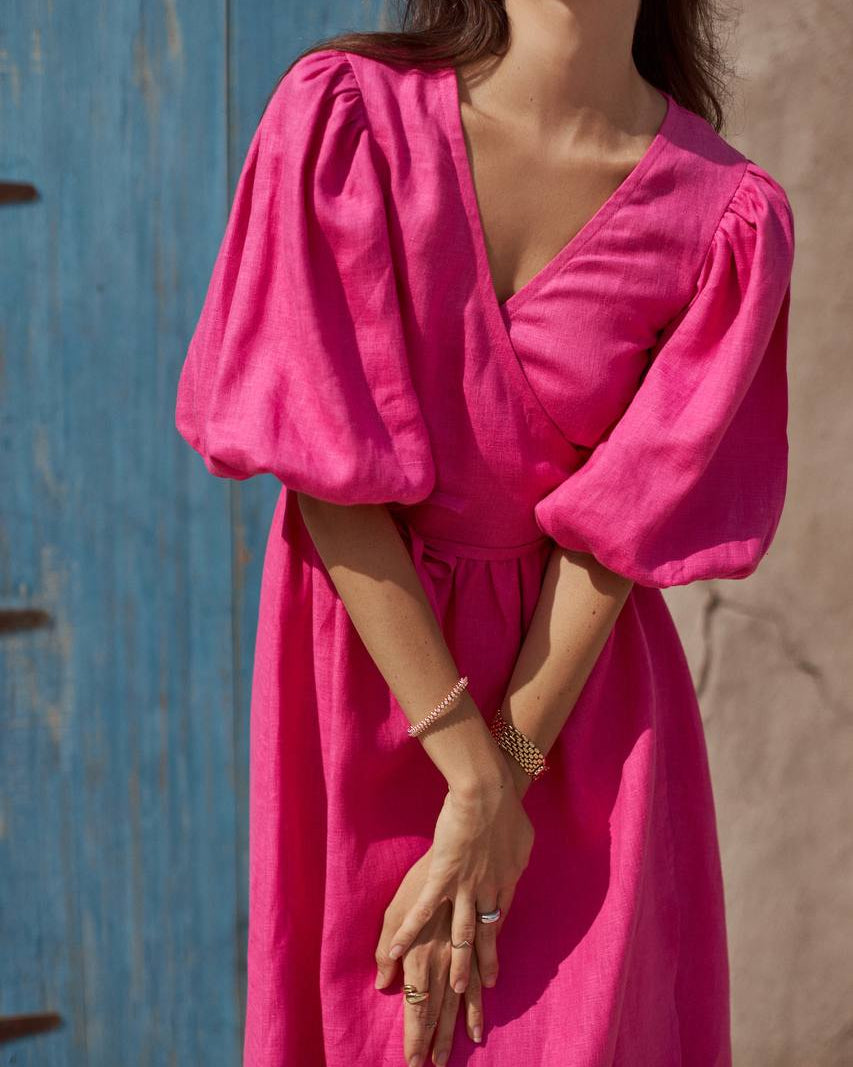 A woman wearing a fuchsia pink midi dress with voluminous puff sleeves and a wrap-style V-neckline, standing against a rustic blue wooden door and beige stucco wall.