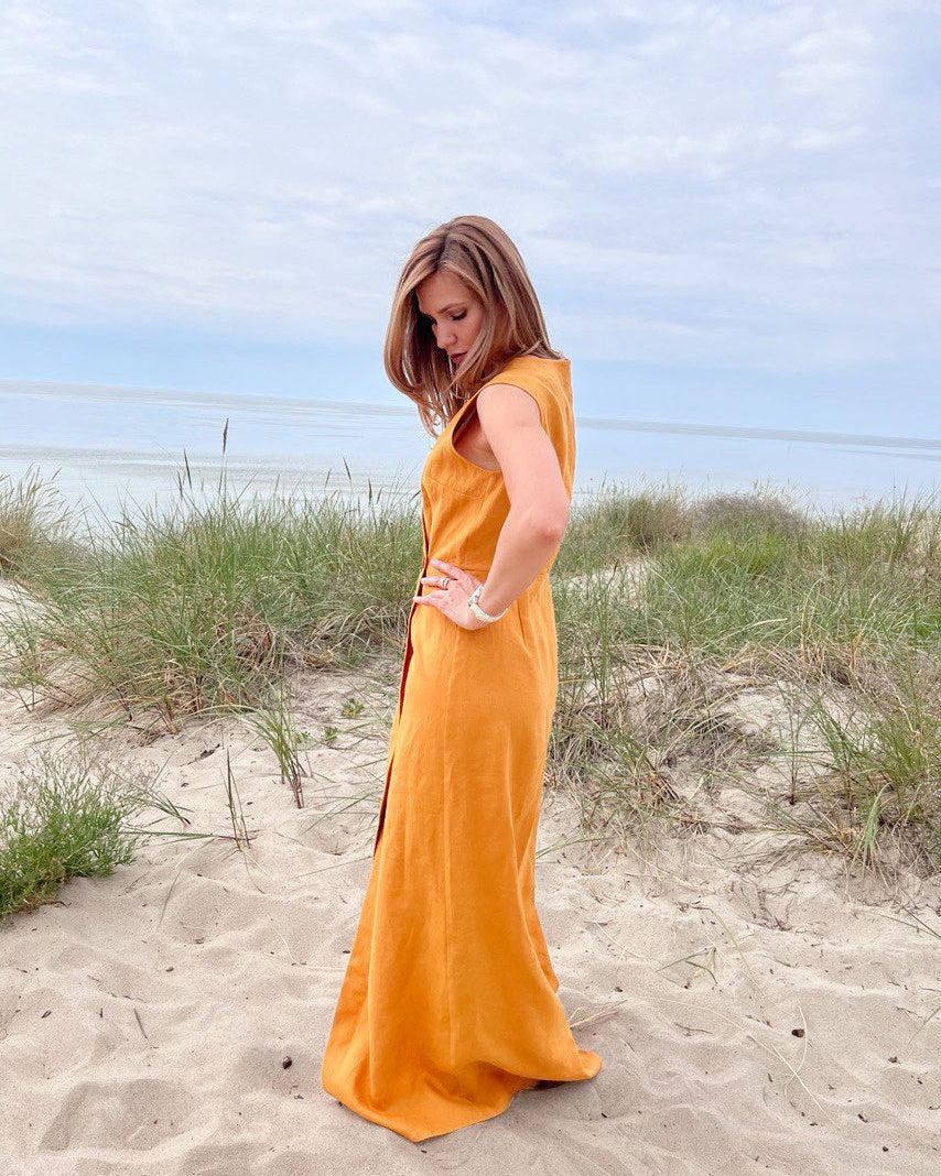 Woman in a mustard dress standing on a sandy beach with grass and blue sky.