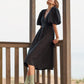 A woman wearing a black puff-sleeve midi dress stands on a wooden deck under a gazebo structure with a beach background.