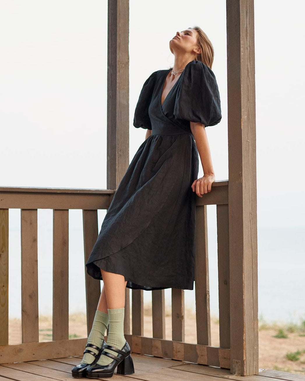A woman wearing a black puff-sleeve midi dress stands on a wooden deck under a gazebo structure with a beach background.