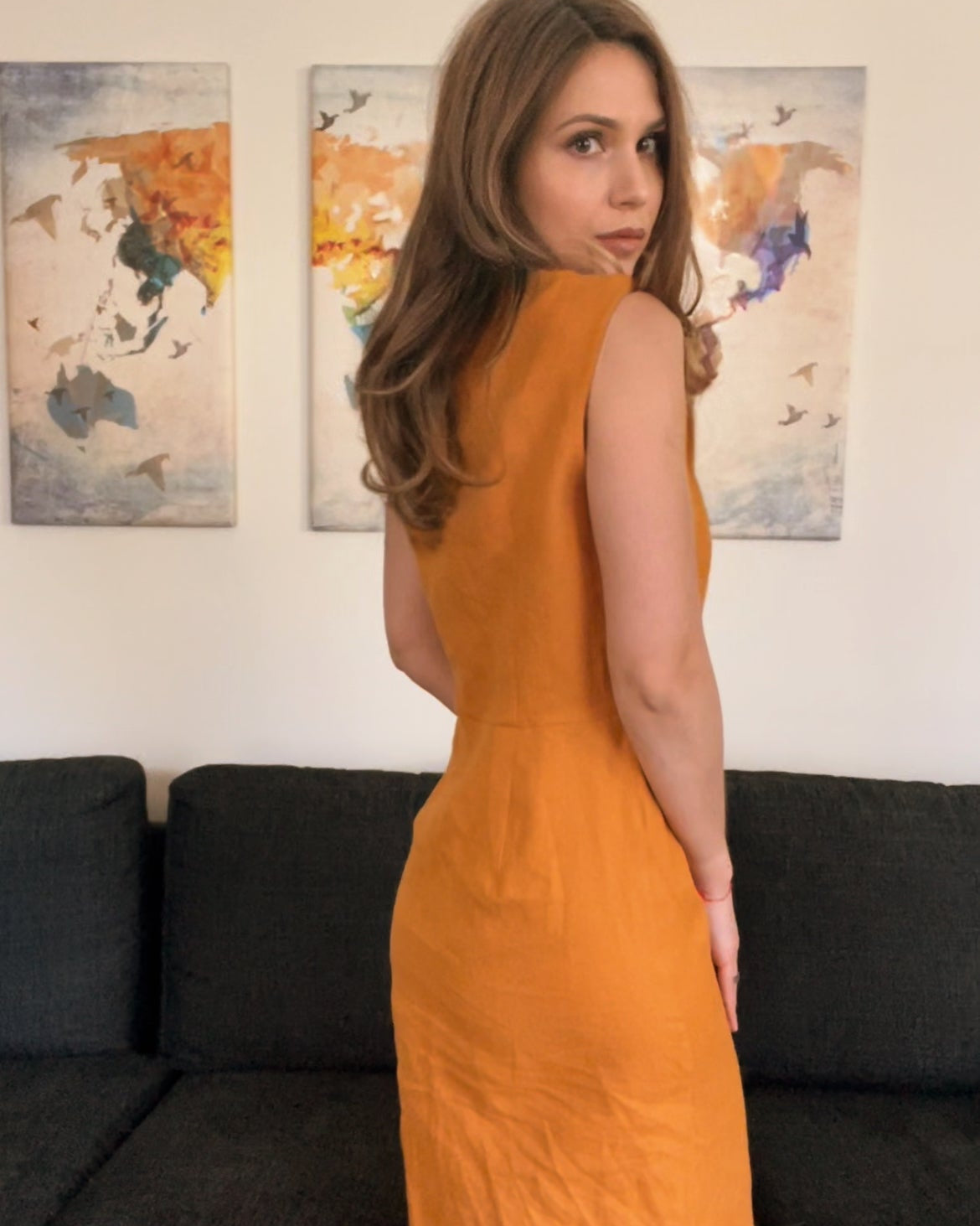 Woman in an orange dress standing in a room with a dark sofa and colorful abstract paintings on the wall.