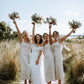 A group of bridesmaids in beige linen dresses with midi length, standing in a field, raising their bouquets in the air.