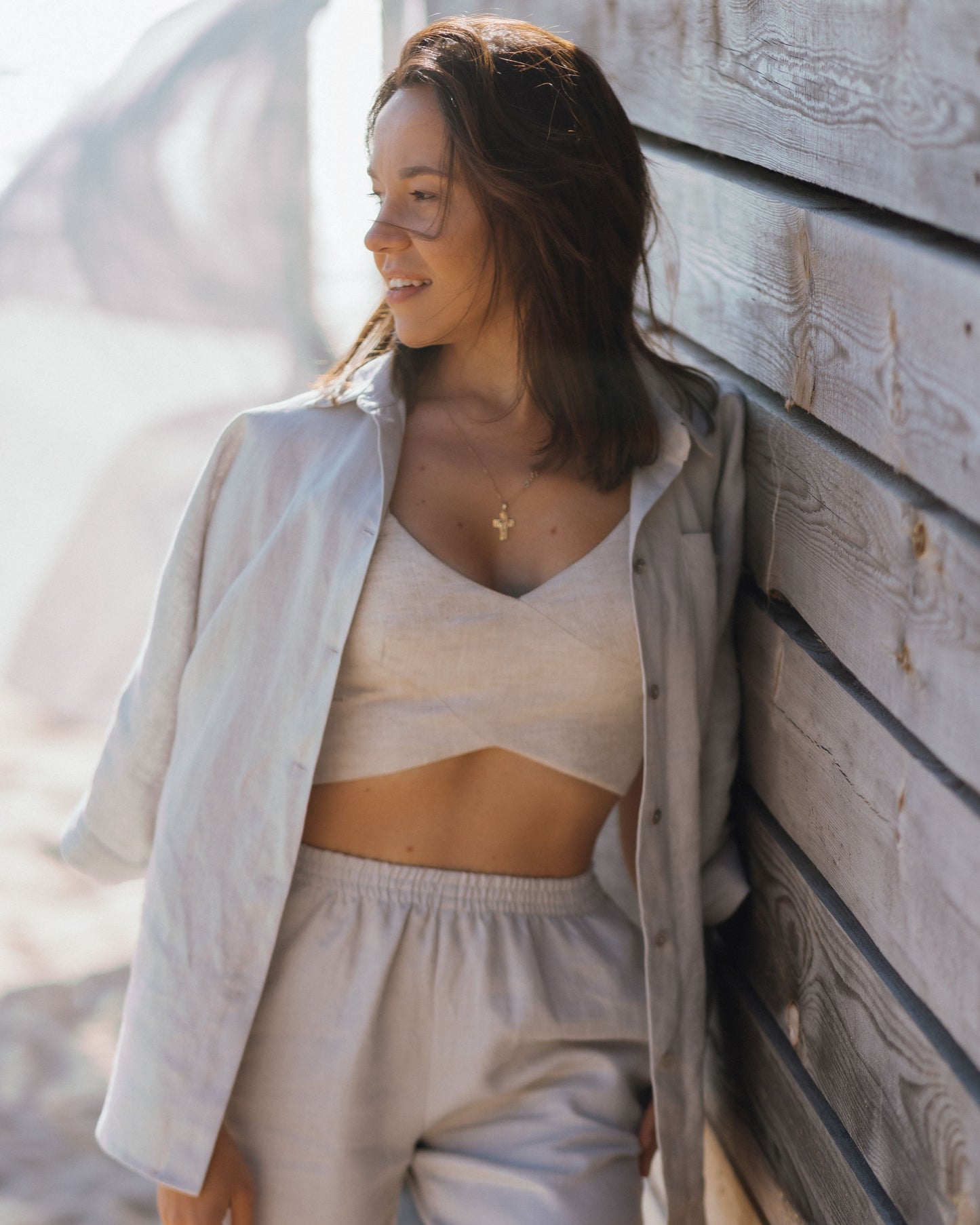 A woman wearing a beige linen crossover cropped top with wide straps and a cross over front detail. She is also wearing light-colored pants and is standing next to a wooden structure.